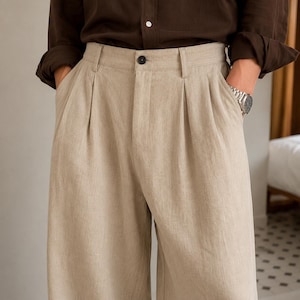 May include: Beige trousers featuring a high-waisted design with front pleats, a central button closure, and belt loops. The pants are paired with a dark brown long-sleeved shirt with a button-down collar and rolled cuffs, creating a neutral ensemble.