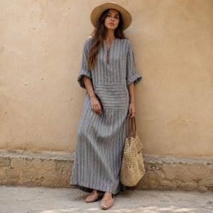 May include: A long-sleeved, floor-length gray dress with vertical stripes, a front pocket, and a small slit at the neckline. The outfit includes a wide-brimmed straw hat, a woven tan tote bag, and light-colored flat shoes, creating a cohesive look.
