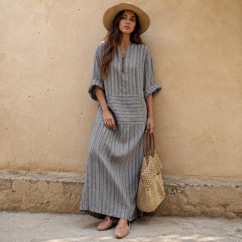 May include: A long-sleeved, floor-length gray dress with vertical stripes, a front pocket, and a small slit at the neckline. The outfit includes a wide-brimmed straw hat, a woven tan tote bag, and light-colored flat shoes, creating a cohesive look.