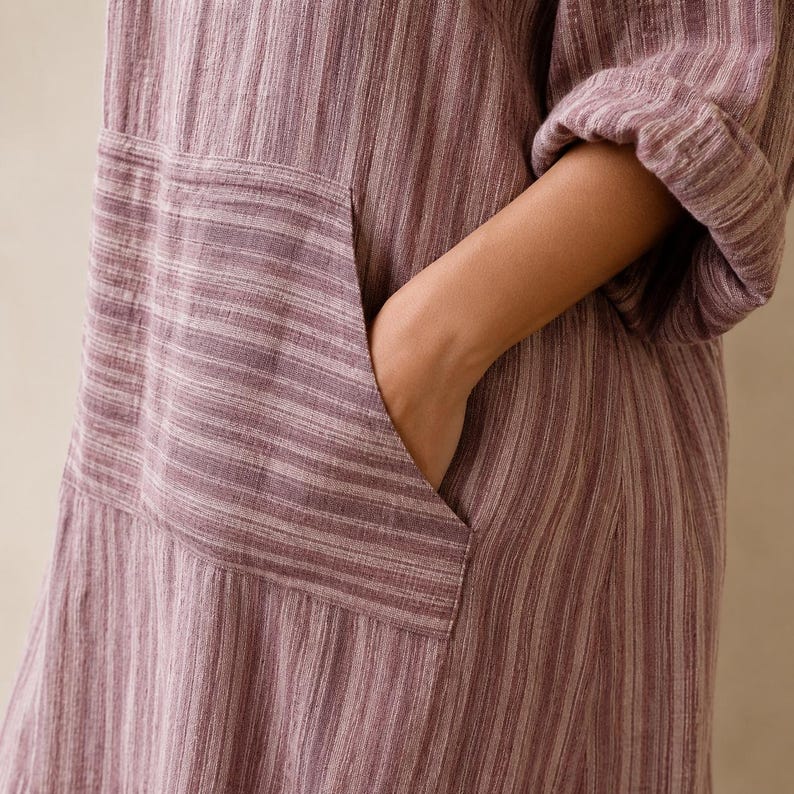 May include: A mauve and white striped dress featuring a textured fabric, long sleeves with rolled cuffs, and a large front patch pocket. The garment displays a vertical striped pattern throughout, with the pocket showcasing a horizontal stripe orientation.