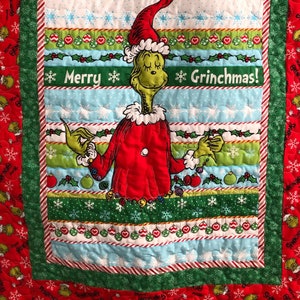 May include: A Christmas-themed quilt featuring the Grinch. The quilt has a red border with Grinch and snowflake patterns. The central panel displays the Grinch in a red Santa suit, with the text "Merry Grinchmas!" in the center. The quilt is made of various colorful fabric strips.