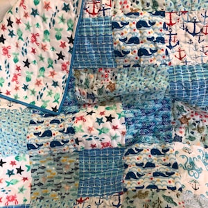 May include: A patchwork quilt with a nautical theme. The quilt is made of various blue, white, and red fabrics with different patterns, including anchors, whales, seashells, and fish.