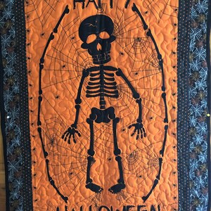 May include: A black and orange Halloween quilt with a skeleton and spiderwebs. The quilt says "Happy Halloween".