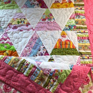 May include: A handmade quilt featuring a patchwork design with white triangles and colorful patterned fabrics. The quilt has a pink polka dot border and a floral patterned backing. The triangles are filled with various patterns, including owls, hearts, and stripes.