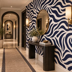 Pode incluir: Um corredor com uma mesa de console de madeira escura com um vaso branco, uma tigela e um livro intitulado The Hotel Book. Acima da mesa, um espelho oval sobre papel de parede com padrão ondulado azul-marinho e branco e duas arandelas.