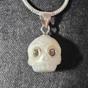 May include: A pendant shaped like a human skull carved from iridescent white mother-of-pearl, featuring two small circular dark stone insets for eyes. The skull is suspended from a silver-toned metal bail attached to a silver-toned snake chain necklace.