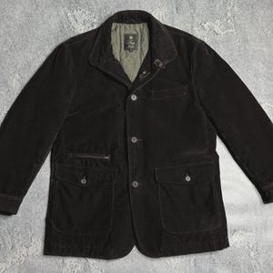 May include: A dark brown corduroy jacket featuring a button-front closure, a pointed collar with a small metal hook-and-eye fastener, two large lower flap pockets, a small zippered chest pocket, and a light olive green quilted interior lining.