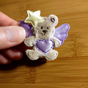 May include: A small, plush teddy bear with purple wings and a heart. The bear has a yellow star on its head and is embellished with purple stitching. The bear is light brown and has a cute, friendly expression.