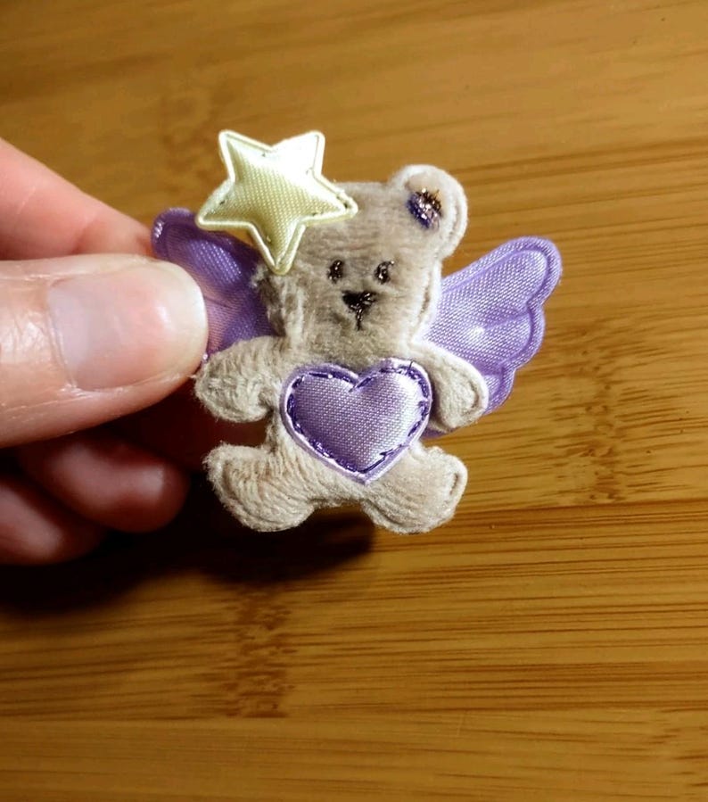 May include: A small, plush teddy bear with purple wings and a heart. The bear has a yellow star on its head and is embellished with purple stitching. The bear is light brown and has a cute, friendly expression.