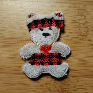 May include: A small, beige, embroidered teddy bear patch featuring a red and black buffalo plaid headband and a matching plaid band around its waist. The bear has a black stitched nose and eyes, and a gold thread necklace with a small red accent.
