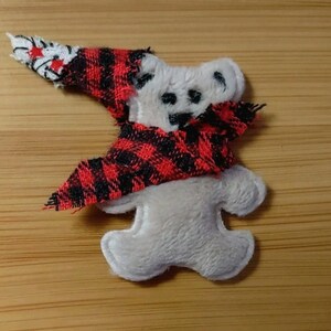 May include: A white plush bear patch featuring a red and black buffalo plaid fabric hat and scarf. The bear has embroidered black eyes and a nose, with the plaid fabric draped over the head and neck area, creating a textured, layered appearance.