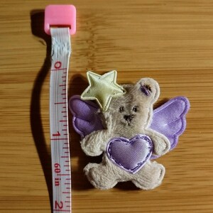 May include: A small, plush teddy bear applique with purple wings and a heart. A yellow star sits atop its head. The applique is approximately 1.5 inches in height. A pink and white measuring tape is next to the applique.