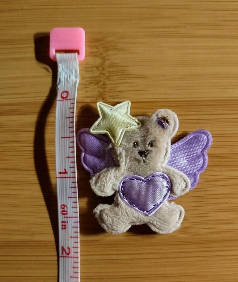 May include: A small, plush teddy bear applique with purple wings and a heart. A yellow star sits atop its head. The applique is approximately 1.5 inches in height. A pink and white measuring tape is next to the applique.