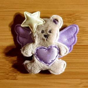 May include: A plush teddy bear with lavender wings and a heart, topped with a cream-colored star. The bear has a stitched black nose and eyes. The heart is lavender with a stitched border. The wings are a matching lavender color.