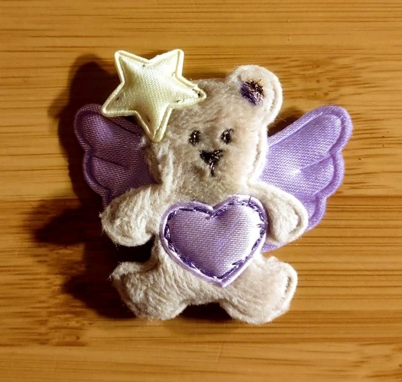 May include: A plush teddy bear with lavender wings and a heart, topped with a cream-colored star. The bear has a stitched black nose and eyes. The heart is lavender with a stitched border. The wings are a matching lavender color.