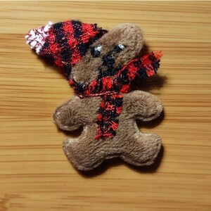 May include: A brown plush gingerbread person applique featuring a red and black buffalo plaid hat and a matching scarf. The figure has embroidered black eyes and a stitched nose, with a soft, fuzzy texture throughout the entire decorative piece.