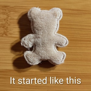 May include: A small, beige teddy bear-shaped felt applique. The bear has a simple design with rounded features and is lying on a wooden surface. The text "It started like this" is at the bottom of the image.