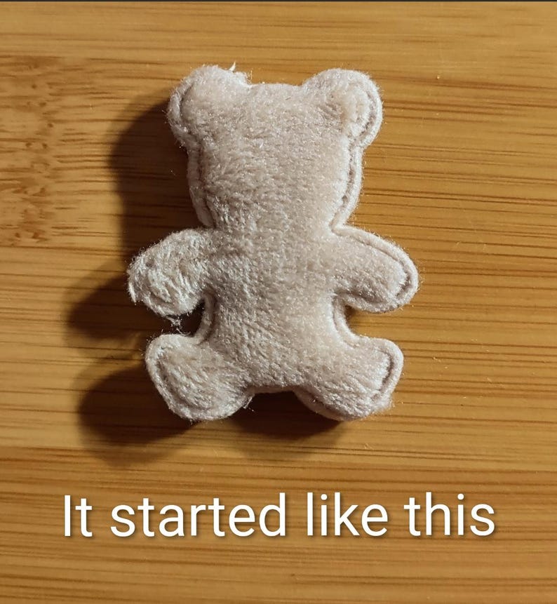 May include: A small, beige teddy bear-shaped felt applique. The bear has a simple design with rounded features and is lying on a wooden surface. The text "It started like this" is at the bottom of the image.