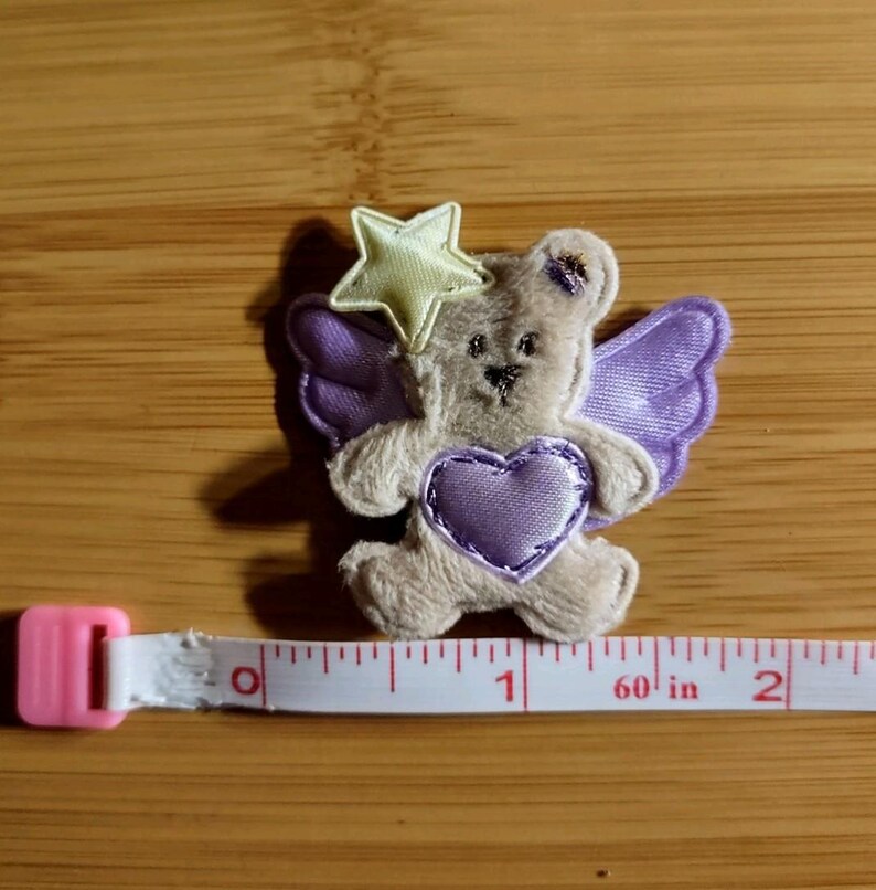 May include: A small, plush teddy bear with purple wings and a heart, and a yellow star above its head. The bear is approximately 1 inch in length, as measured by a pink and white measuring tape.