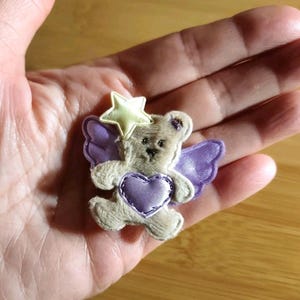 May include: A small, plush teddy bear with purple wings and a heart, and a yellow star on its head. The bear is light brown with a purple heart and wings. The star is yellow with a white outline. The overall design is whimsical and cute.