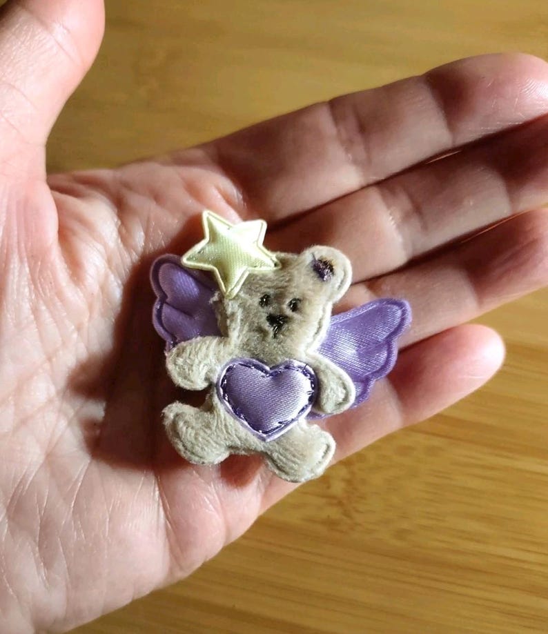 May include: A small, plush teddy bear with purple wings and a heart, and a yellow star on its head. The bear is light brown with a purple heart and wings. The star is yellow with a white outline. The overall design is whimsical and cute.