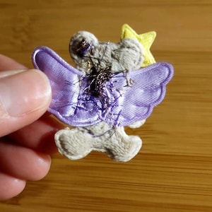 May include: A fabric angel applique with a gray body, lavender wings, and a yellow star. The angel has metallic thread accents and is held by a hand against a wooden background. The wings have a scalloped edge.