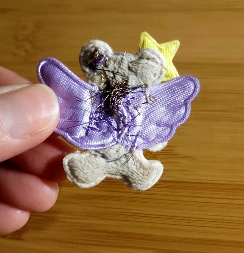 May include: A fabric angel applique with a gray body, lavender wings, and a yellow star. The angel has metallic thread accents and is held by a hand against a wooden background. The wings have a scalloped edge.