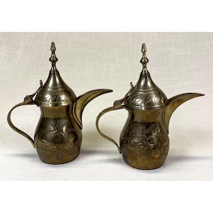 May include: Two brass coffee pots with pointed lids, curved handles, and long spouts. The left pot features an engraved palm tree and crossed swords, while the right pot displays an engraved floral pattern. Both have a metallic finish.
