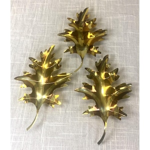 May include: Three gold-colored metal leaf-shaped dishes with a polished, reflective surface. Each dish features a detailed, multi-lobed oak leaf silhouette with a pointed stem at the base, arranged in a cluster on a light-colored fabric surface.