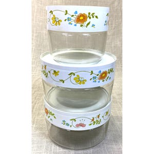 May include: A stack of three clear glass storage bowls with white lids. Each lid features a decorative floral pattern with orange, yellow, and blue flowers on green vines. The bowls are cylindrical with smooth sides and transparent glass bases.