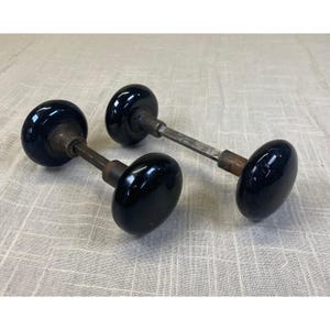 May include: Two matching door knobs with smooth, glossy black spherical handles. Each knob is connected by a square metal spindle and features a weathered, dark metallic base where the handle meets the central connecting rod.
