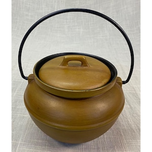 May include: A round, mustard-yellow ceramic pot with a matching lid featuring a small handle. The pot has a black metal swing handle attached to two side lugs. The interior rim of the pot is black, and the exterior has a smooth, matte finish.