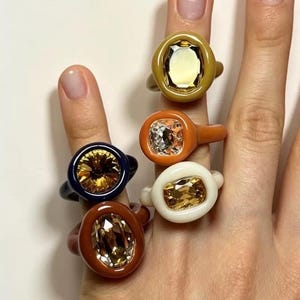 以下が含まれることがあります： マスタードイエロー、バーントオレンジ、ネイビーブルー、テラコッタ、クリーム色の滑らかな無地の樹脂バンドを備えた5つのチャンキーリングのコレクション。各リングの中央には、透明または琥珀色の大きなファセットカットの宝石がセットされています。