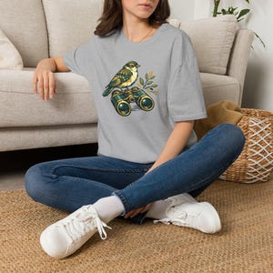 Könnte beinhalten: Ein meliertes graues T-Shirt mit einer detaillierten Illustration eines Vogels, der auf einem Fernglas sitzt. Der Vogel hat grüne, gelbe und braune Federn, und das Fernglas hat goldene Akzente. Das Design beinhaltet Olivenzweige. Ein lässiges T-Shirt zum Thema Natur.