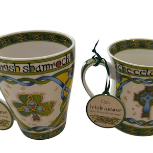 Könnte beinhalten: Zwei weiße Tassen aus Knochenporzellan mit grünen und blauen keltischen Knotenmustern. Die linke Tasse zeigt ein grünes Kleeblatt und den Text "the irish shamrock". Die rechte Tasse zeigt ein keltisches Kreuz und den Text "the celtic cross". Beide Tassen haben grüne Schnuretiketten mit der Aufschrift "Clara Irish Weave new bone china mug".