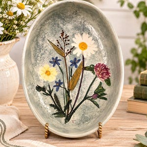 May include: An oval ceramic dish with a mottled light blue and grey glaze, featuring a pressed botanical arrangement of a white daisy, a yellow flower, small blue blossoms, a dark pink clover, and green leaves centered on the interior surface.