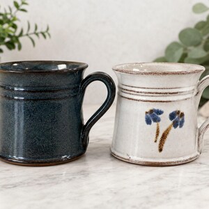 May include: Two ceramic mugs with a flared rim and a sturdy handle. One mug features a deep, speckled navy blue glaze, while the other is a creamy off-white with a hand-painted blue and brown floral motif on the front side of the vessel.