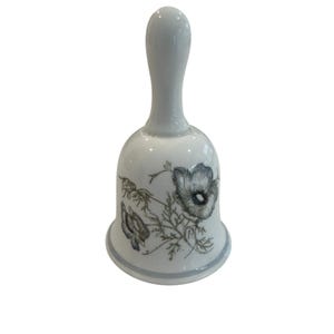May include: A white porcelain bell featuring a slender handle and a flared base. The exterior surface is decorated with a detailed botanical illustration of a dark grey flower with delicate petals and thin stems, finished with a light grey rim.