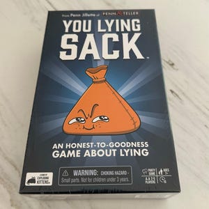 May include: A rectangular board game box titled You Lying Sack from Penn Jillette of Penn & Teller. The dark blue box features a central orange sack character with a mischievous face, and the text An Honest-to-Goodness Game About Lying.