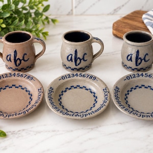 May include: Three ceramic mugs and three matching saucers in tan and grey tones. Each mug features the blue text abc and a handle. Each saucer displays the blue text 0123456789 and the alphabet in a circular pattern with a blue checkered border.