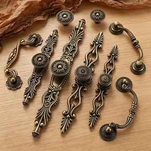 May include: An assortment of antique brass cabinet hardware, including knobs and pulls. The hardware features intricate floral and scrollwork designs. The knobs and pulls are in various sizes and shapes, suitable for drawers and cabinets. The hardware is displayed on a light wood surface.
