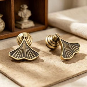 May include: Two brass-toned metal cabinet pulls shaped like ginkgo leaves. Each pull features a circular base with concentric rings and a fan-shaped handle with dark, recessed grooves radiating from the center to the scalloped outer edge.