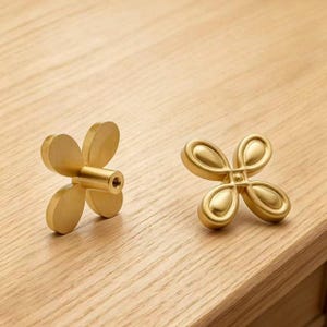 Puede incluir: Dos pomos de armario de metal color latón con forma de flor de cuatro pétalos. Un pomo se muestra de lado, revelando un poste de montaje cilíndrico con un orificio roscado, mientras que el otro se muestra de frente, con un diseño de pétalos en relieve.