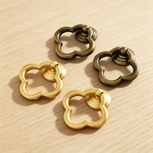 May include: Four metal cabinet pulls with a clover-shaped ring design. Two pulls feature a brushed brass finish, while the other two have an antique bronze finish. Each pull includes a circular mounting base and a hinged ring for gripping.