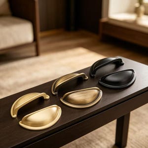 May include: A collection of six cabinet pulls in three distinct finishes: matte gold, brushed brass, and matte black. The set includes three arched bar-style handles and three semi-circular cup-style pulls, all arranged on a dark wooden surface.