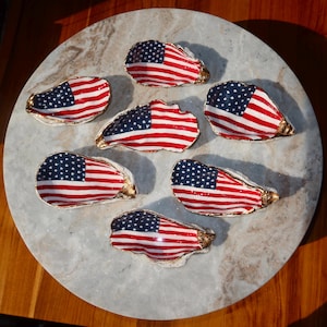May include: Six oyster shells arranged in a circular pattern, each painted with the American flag featuring red and white stripes and a blue field with white stars. The edges of each shell are finished with a metallic gold trim.