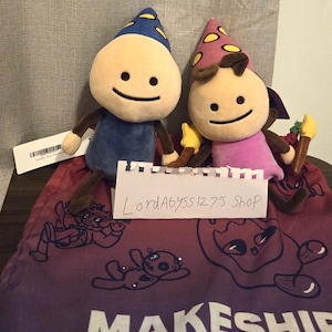 Puede incluir: Dos muñecos de peluche con caras redondas de color beige y rasgos de líneas negras simples. Uno lleva un sombrero azul con estrellas amarillas y cuerpo azul, el otro un sombrero rosa con círculos amarillos y cuerpo rosa sobre una bolsa morada MAKESHIP.