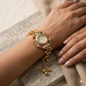 May include: Gold-toned watch with a floral design, featuring red, green, and white accents. The watch face is white with black numerals. The watch is attached to a chain with small gold bells. The brand name "VELMORA BY MYRA" is visible.