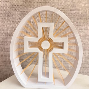 May include: A white, oval-shaped decorative frame containing a central white cross. Thin, golden-colored radial lines extend from the center of the cross to the inner edge of the oval frame, creating a sunburst effect within the structure.