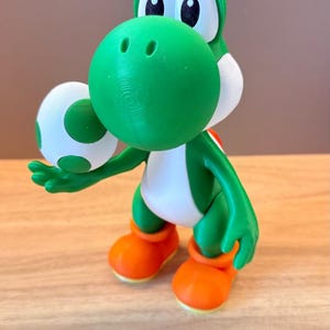 May include: A green and white Yoshi figurine holding a white egg with green spots. The character has large white eyes with black pupils, a white belly, and bright orange shoes. The figure is constructed with visible 3D printed layer lines.